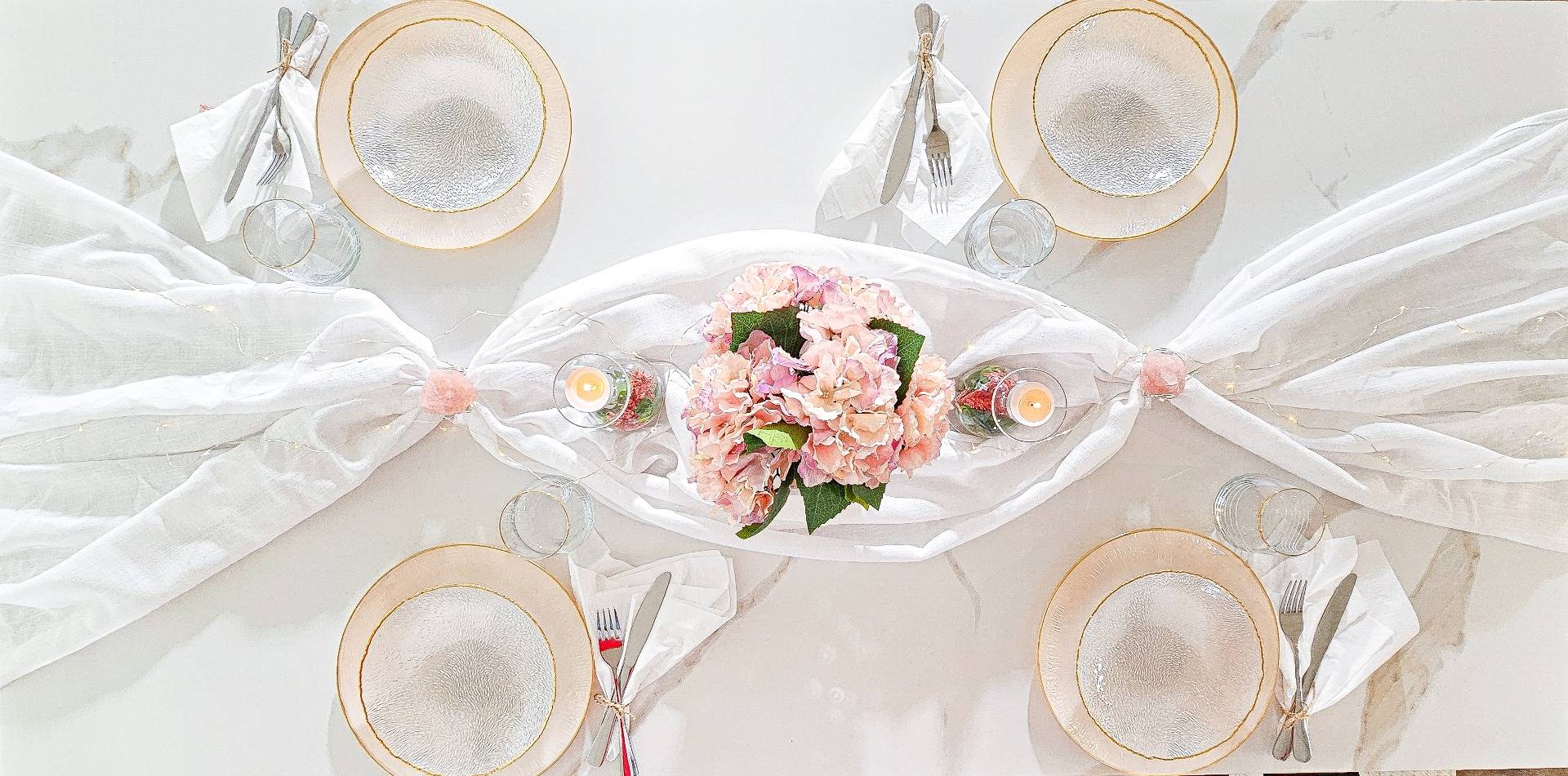 Bild på ett uppdukat bord till middagsbjudning. På bilden ser man tallrikar, ljus, blommor, duk, servettringar, bestick och glas. Bilden är tagen ovanifrån